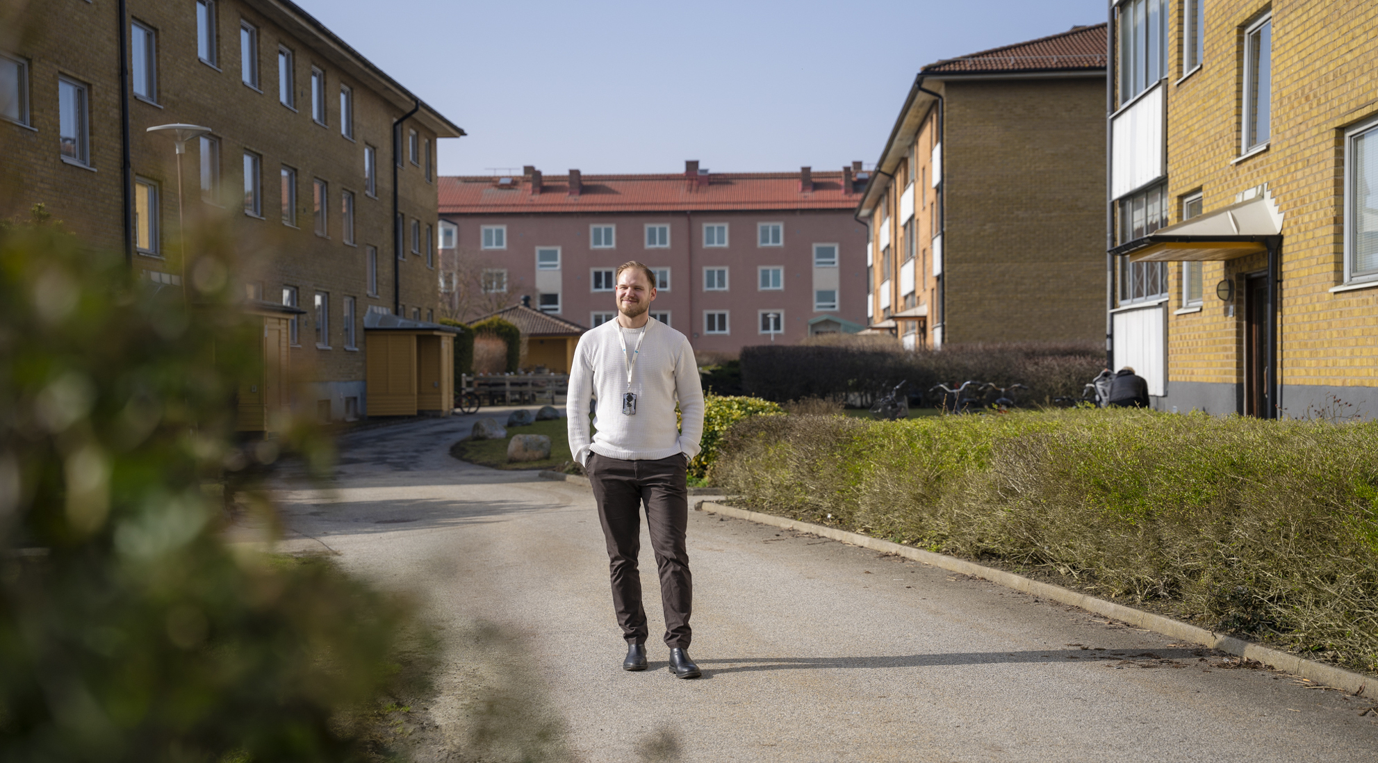 En man med vit tröja och bruna byxor som står på en grönskandea gård i ett bostadsområde